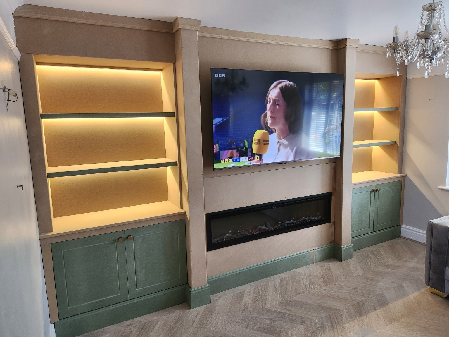 Media wall during installation with unpainted MDF, green cupboard fronts, recessed TV and warm shelf lighting