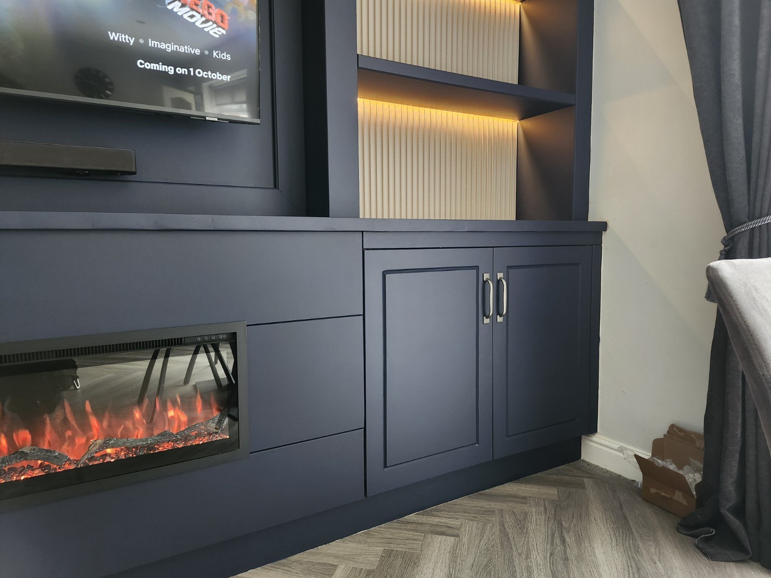 Close view of navy panelled cupboard doors, silver handles, electric fire and illuminated shelving