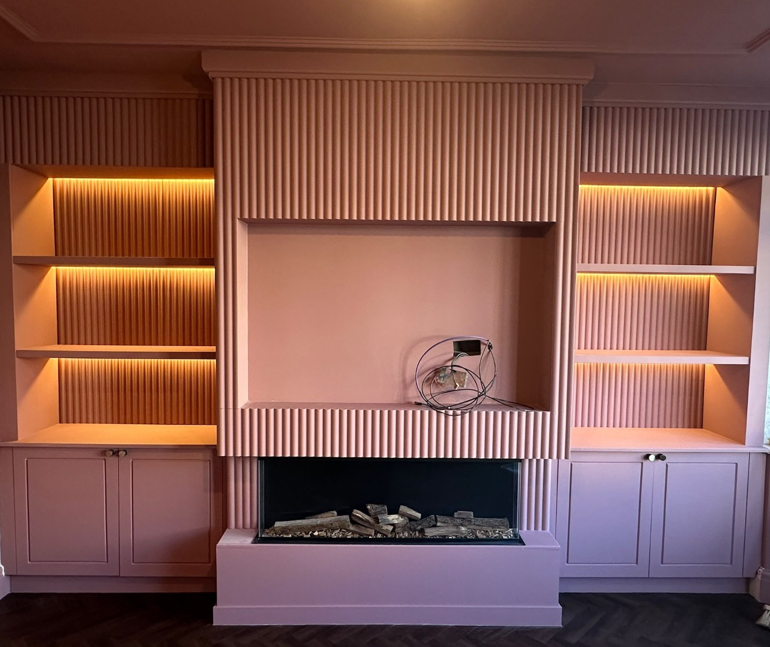 Painted media wall in a dusky pink finish, with warm LED shelf lighting, fitted cupboards and a recess ready for the TV