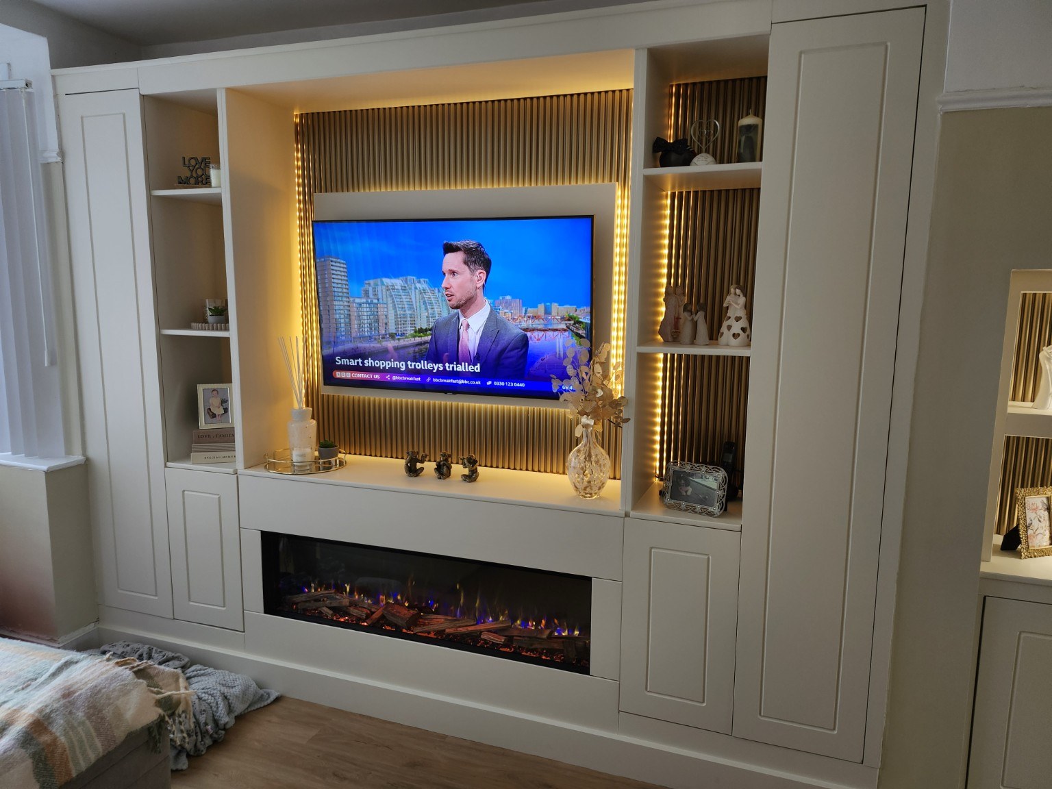 White media wall with tall storage cupboards, fluted TV backing, warm lighting and a wide electric fire